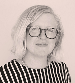 headshot photo of a white woman with bleached blond hair and rimmed glasses wearing a black and white striped shirt